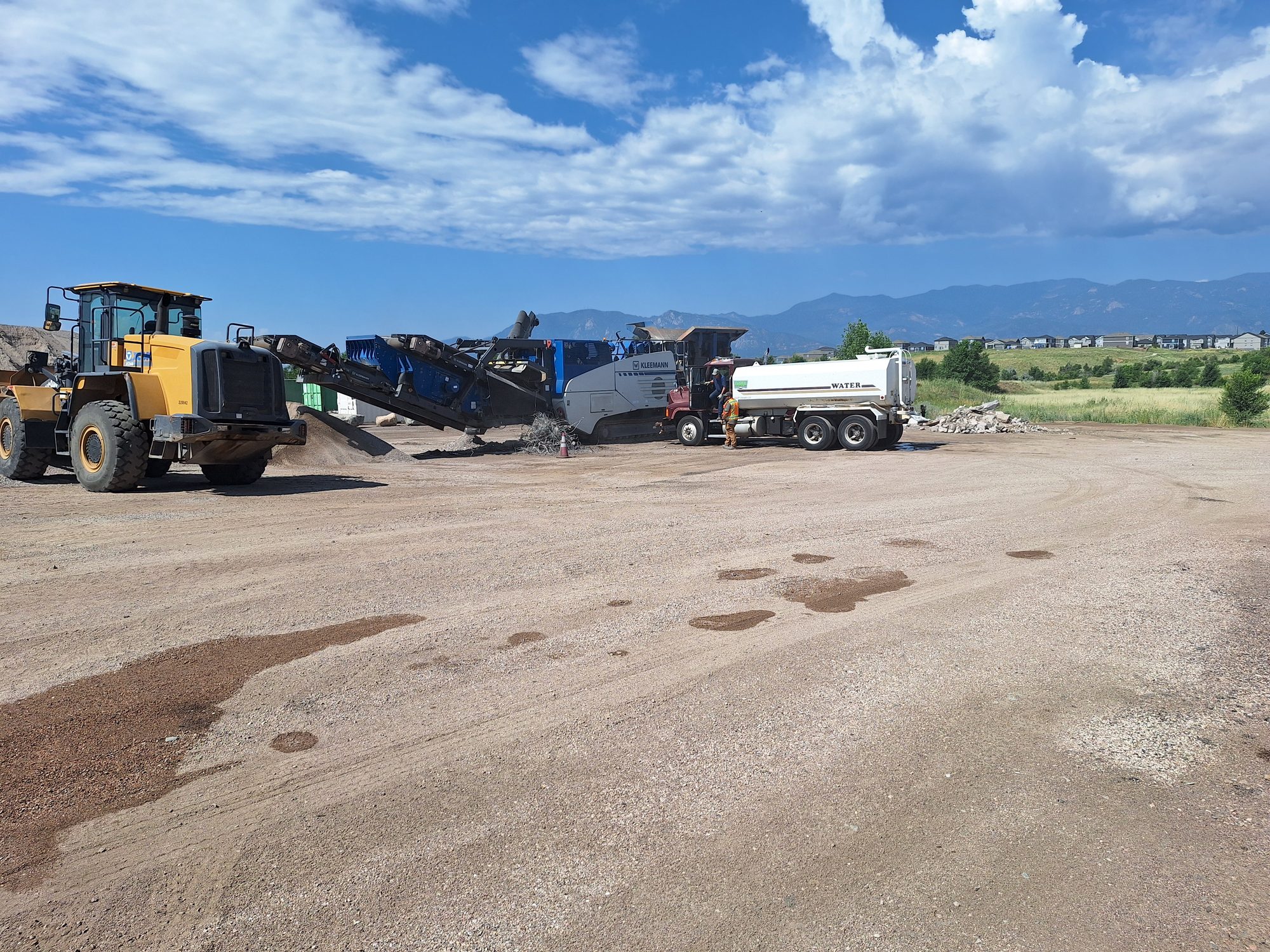 CrushRite equipment lineup — Kleemann screen plant, water truck, and wheel loader operating with Pikes Peak in the background